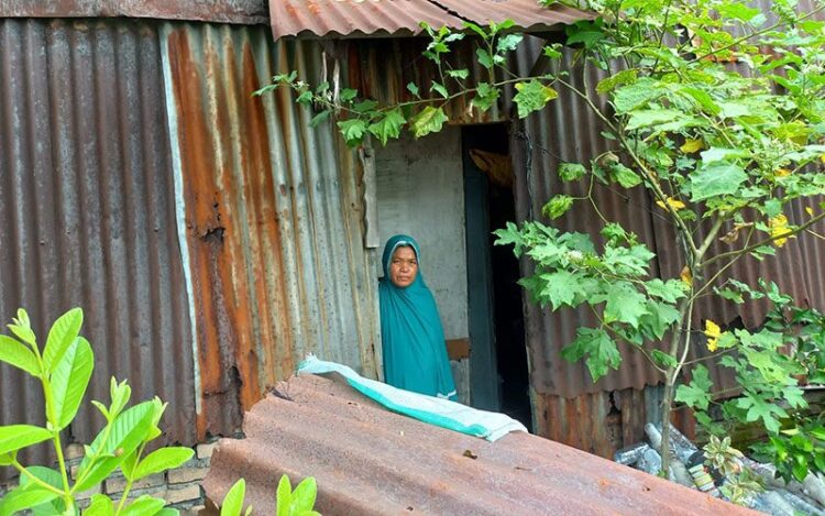 Syamsinar berdiri di depan rumahnya yang terbalut seng bekas dan berkarat. Sekitar rumahnya juga terbalut semak dan tumpukan botol-botol minuman bekas.
